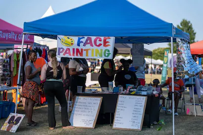 People gather around an outdoor face painting booth with a 'Face Painting' sign and colorful designs.