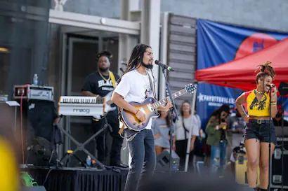 A male guitarist with dreadlocks sings into a microphone with a female singer and keyboardist behind him, all performing outdoors.