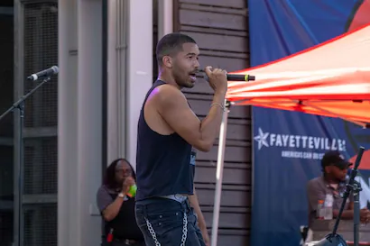 A man in a black tank top and jeans with chains sings into a microphone on an outdoor stage.