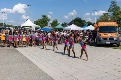 Five young girls in pink and blue perform on an outdoor paved area, with a crowd and tents in the background.