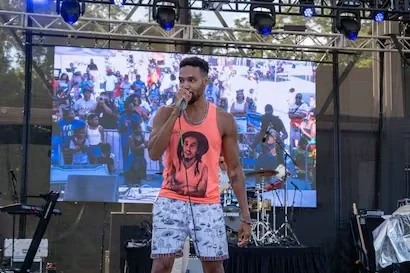 A man in an orange Bob Marley tank top sings into a microphone on an outdoor stage. A screen behind him shows a crowd.